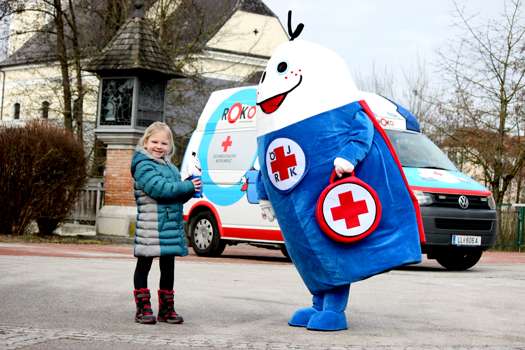 ROKO mit dem roten Herz, das Maskottchen vom JRK, steht mit einem blonden Mädchen vor dem Roko-Mobil.