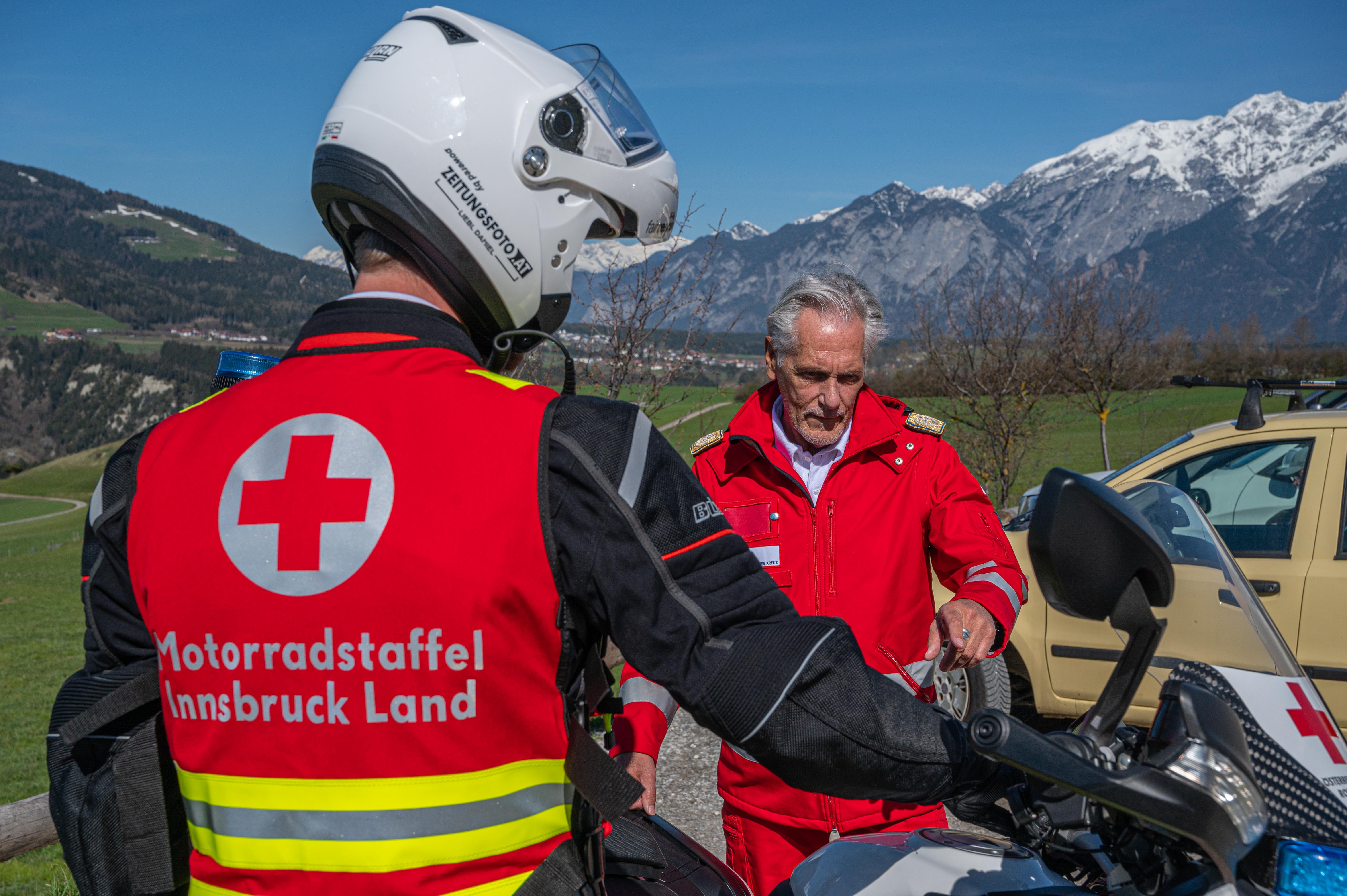 Jan Reiter, in seiner Motorrad-Einsatzuniform steht vor seinem Einsatzmotorrad und erklärt Rotkreuz-Präsident Günther Ennemoser in Rotkreuz-Uniform die Funktionsweise des Motorrades, das alles vor der Tiroler Bergkulisse